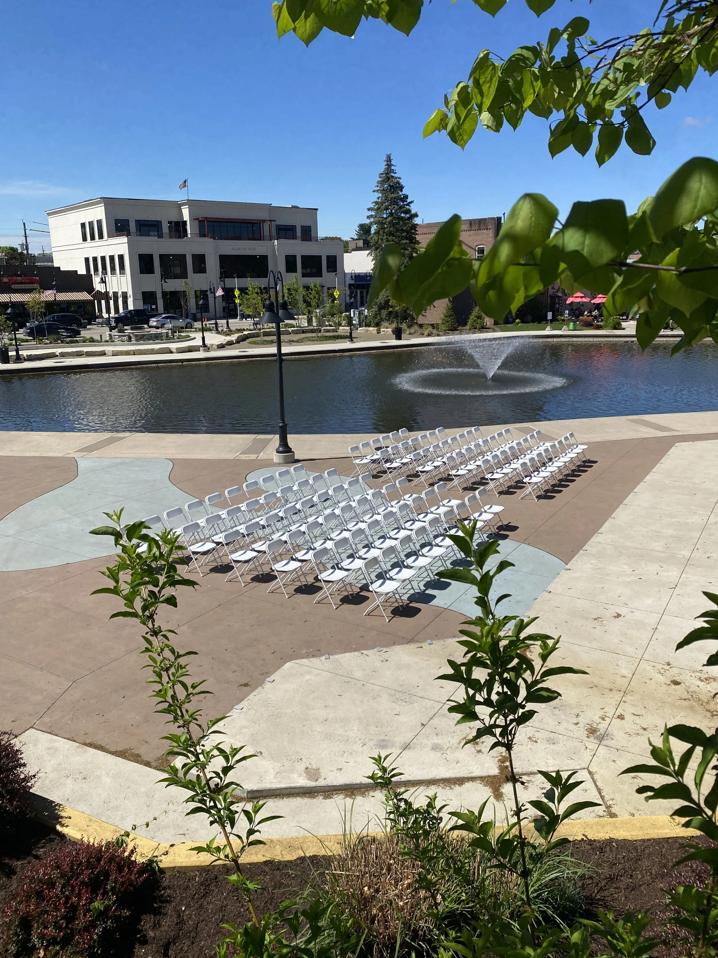 Outdoor wedding ceremony at the Mill Pond in Brighton, Michigan with white rental chairs beside the pond and fountain.