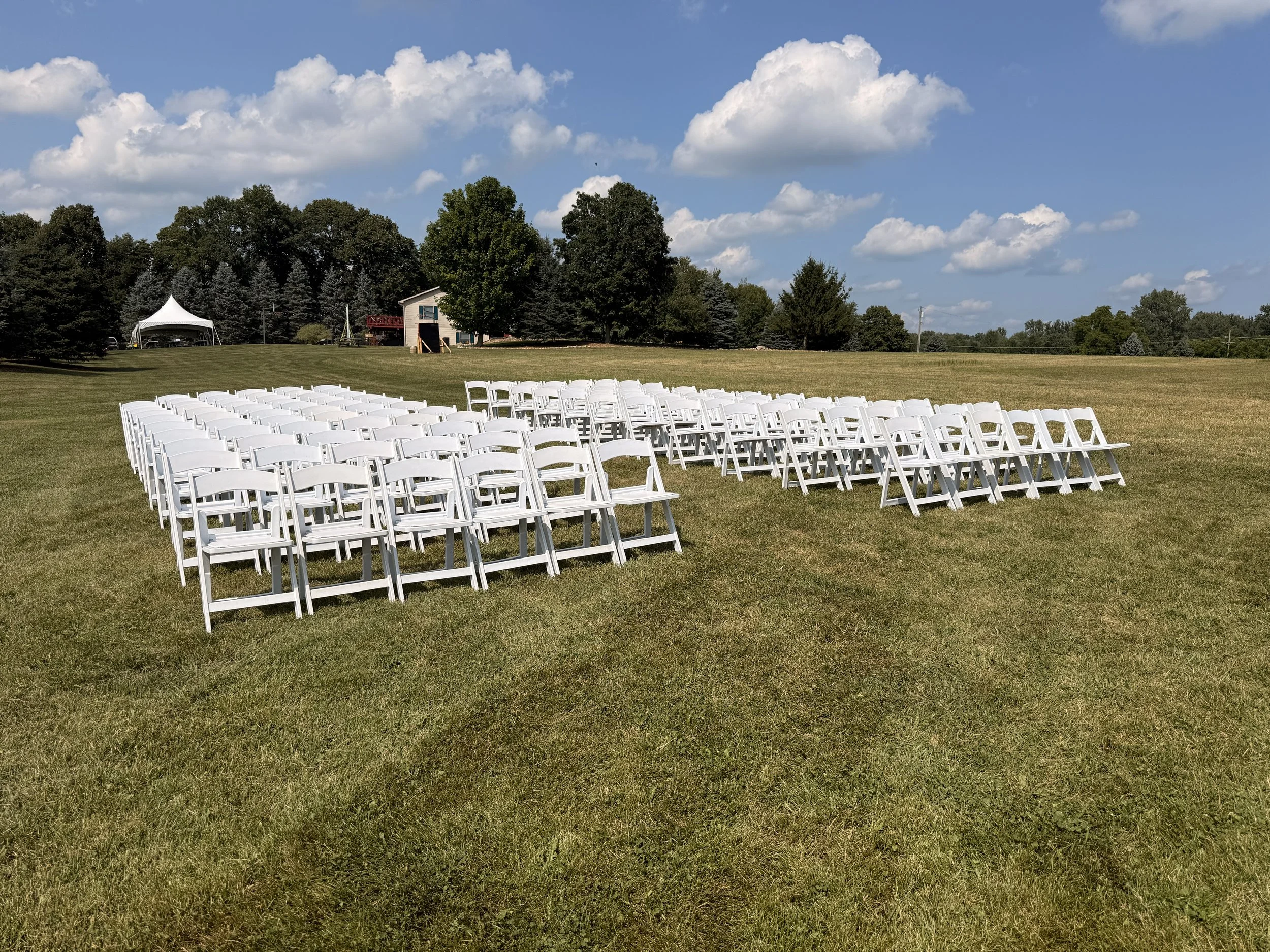 130 white padded resin chairs set up for an outdoor wedding ceremony.