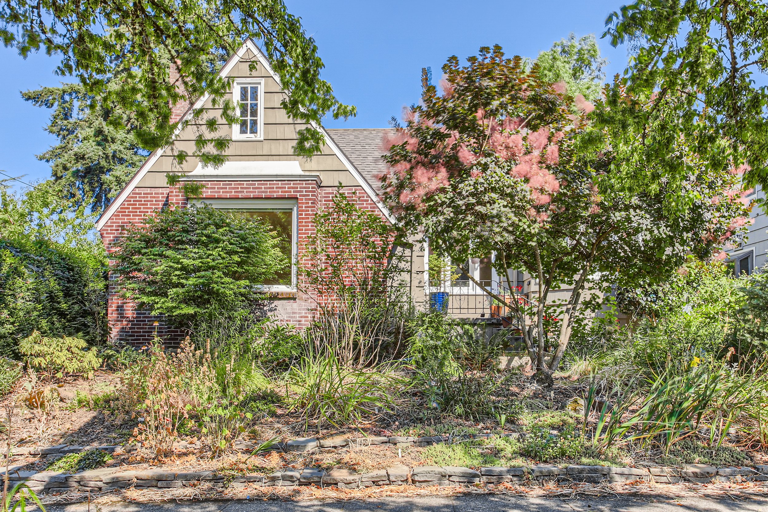 Front view of a brick house in Portland, OR with a triangular roof, surrounded by trees and plants.
