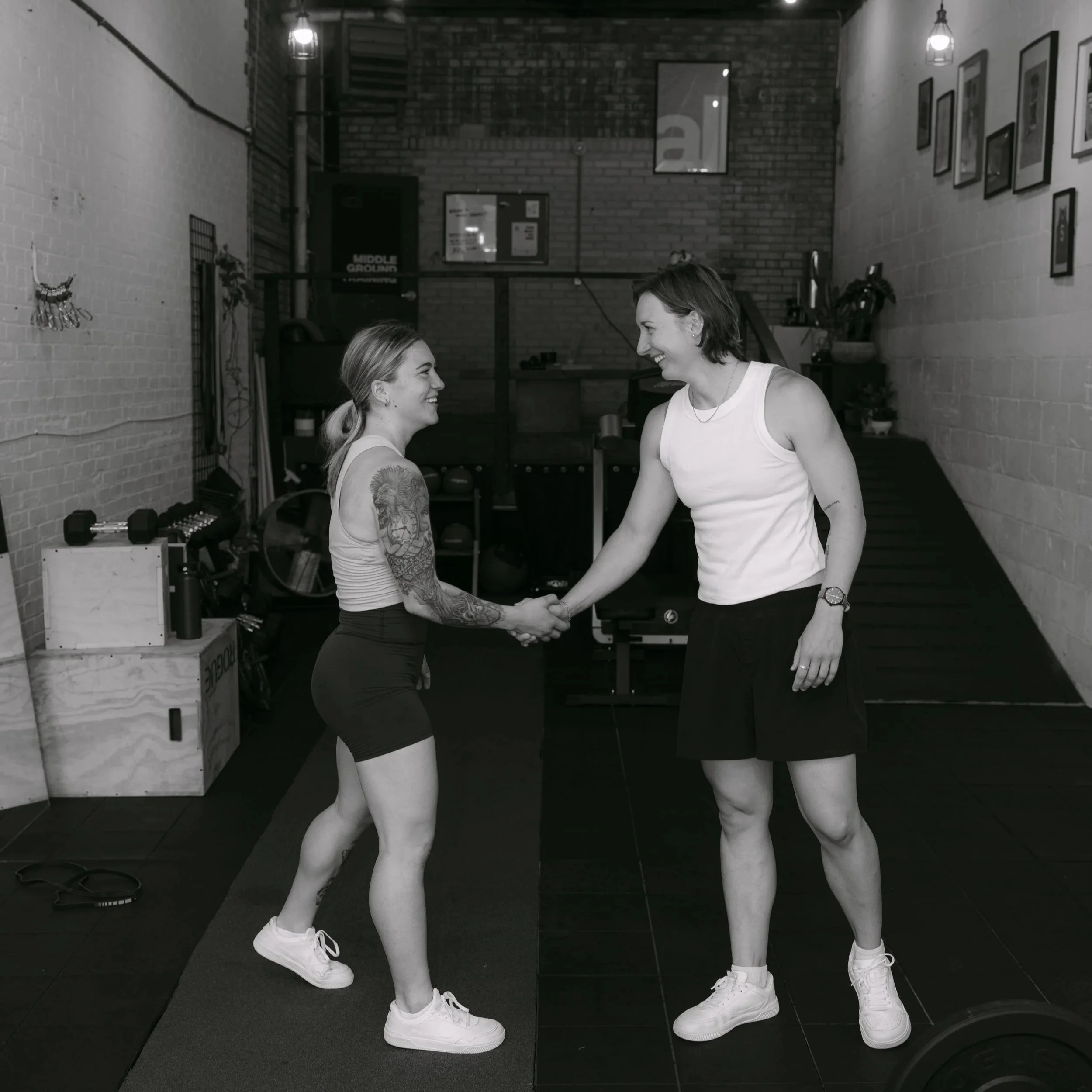 Two women are smiling and holding hands in a gym setting, both dressed in athletic wear with sneakers, standing on a black floor in front of a brick wall with framed pictures and hanging lights.