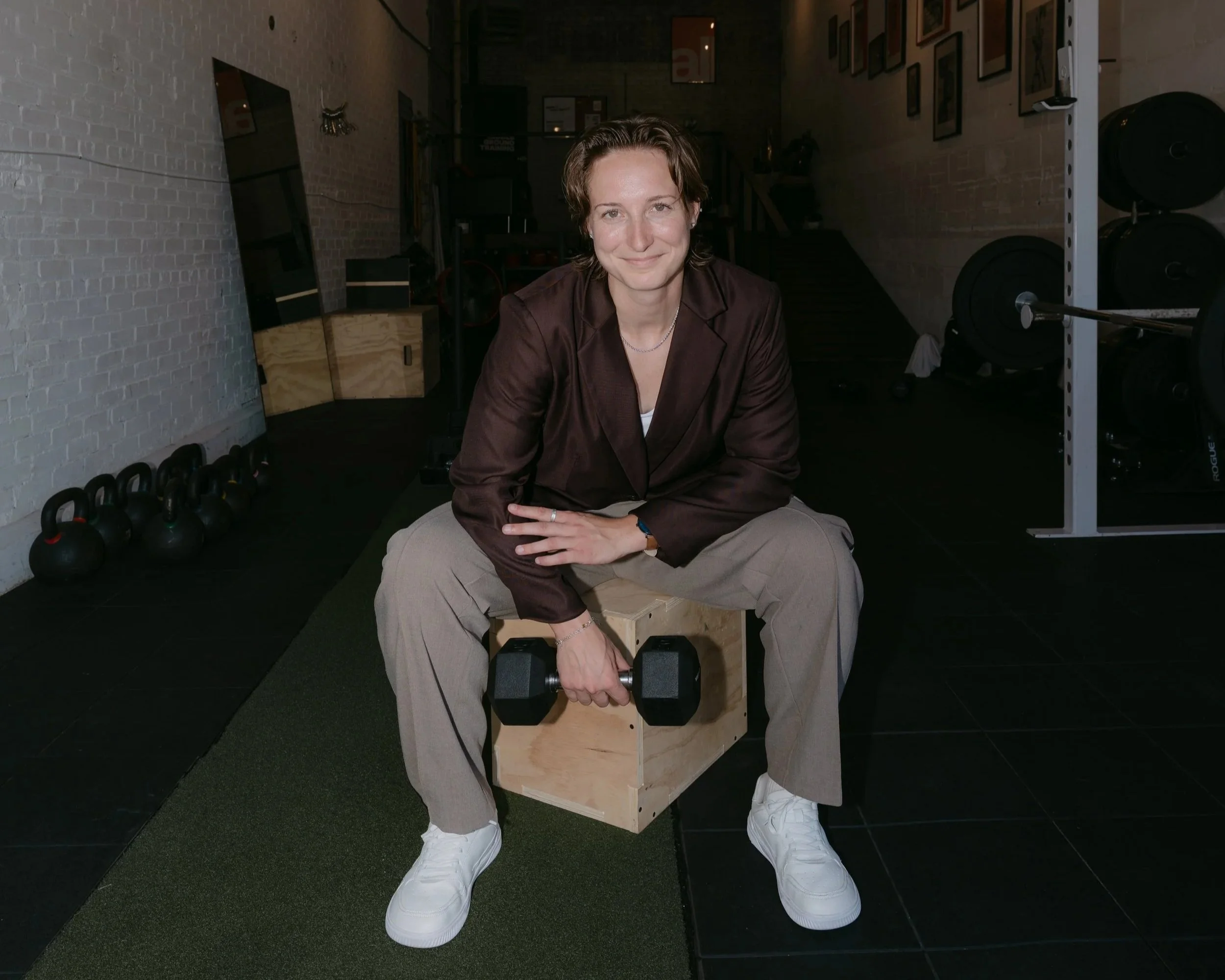 A smiling woman seated on a wooden box, holding a dumbbell in her right hand, in a gym with kettlebells on the floor to her left, weight plates behind her and framed pictures on the wall to her right.