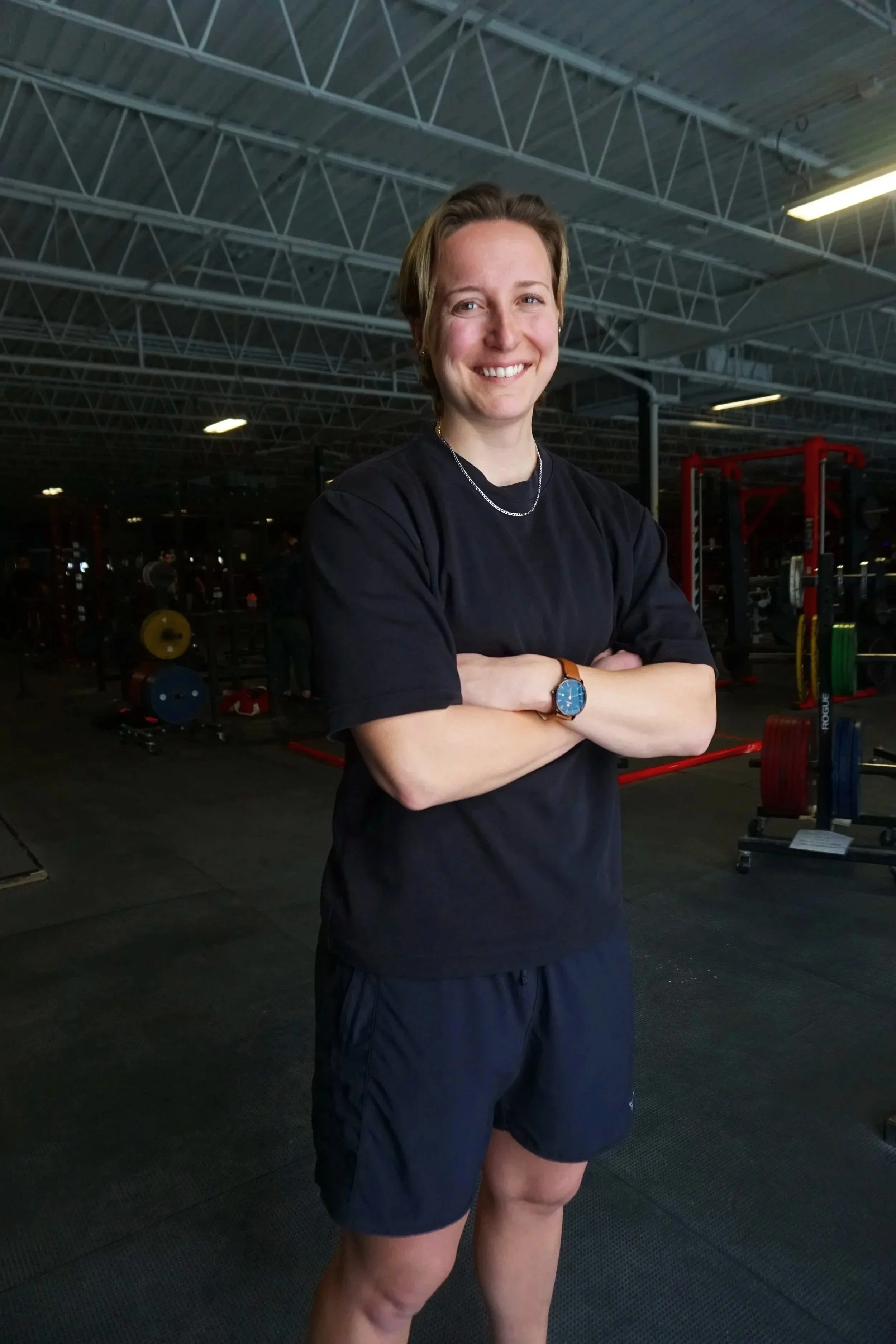 A smiling woman with short hair in a gym, wearing a black t-shirt, black shorts, a watch, and a necklace, standing with arms crossed.