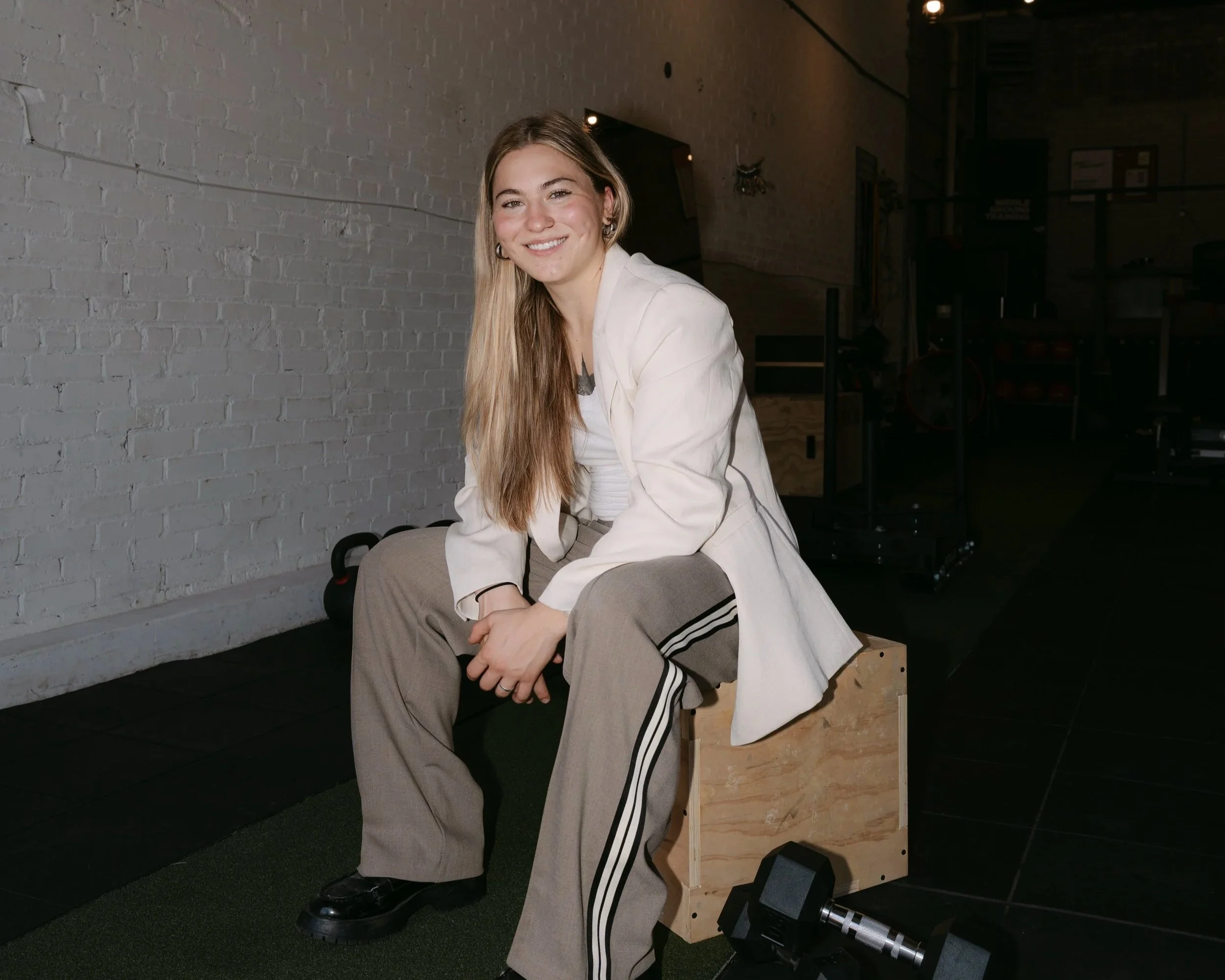 A young woman with long blonde hair, wearing a white jacket and gray sweatpants with black stripes, sitting on a wooden exercise box in a gym. She is smiling and looking at the camera. The background includes a white brick wall, some gym equipment, and a black floor with green turf.