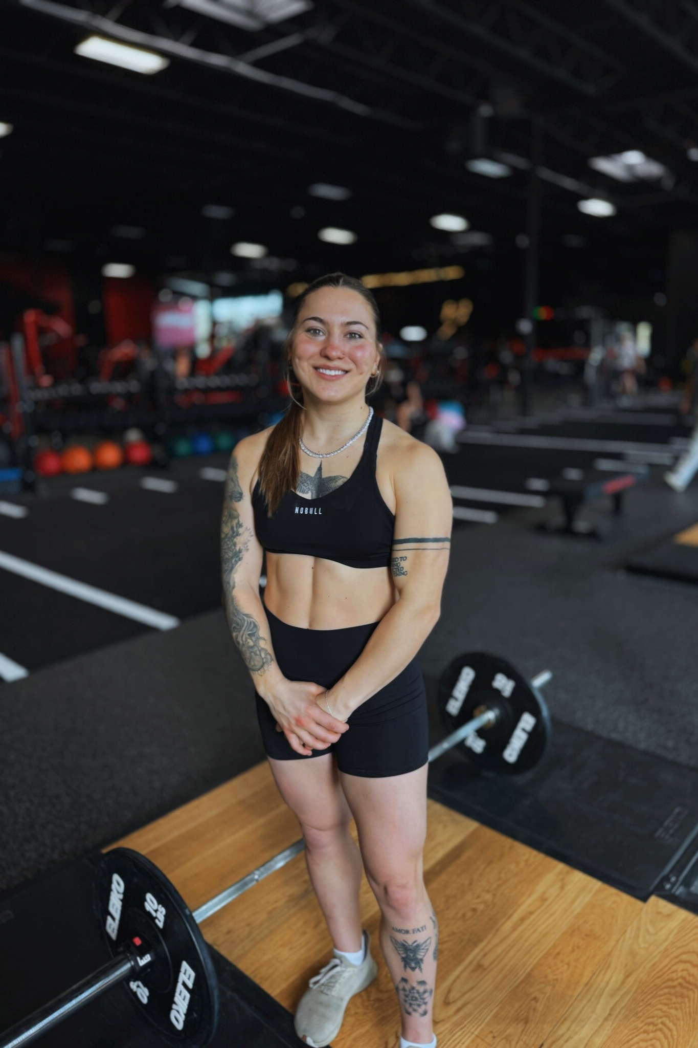 Woman in athletic wear standing in a gym with weights and workout equipment in the background.