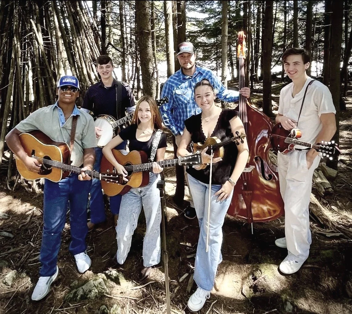 A group of six young musicians with guitars, a violin, a banjo, and a double bass standing in a forest with trees and sunlight filtering through.