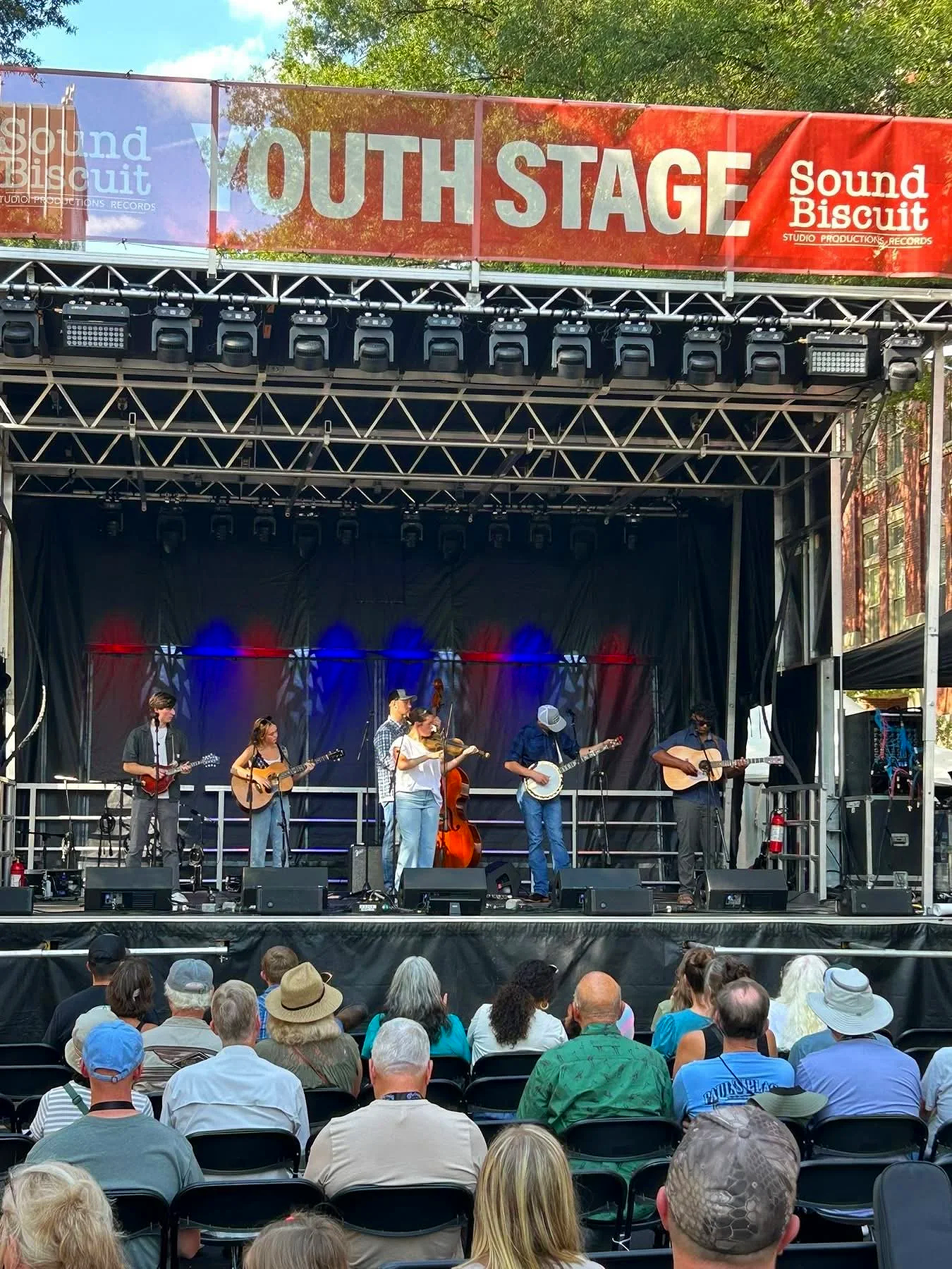 A band of seven musicians performs on an outdoor stage at a music festival. The stage has a red banner reading 'YOUTH STAGE' and 'Sound Biscuit.' The audience includes a diverse group of people sitting in chairs, watching the show.