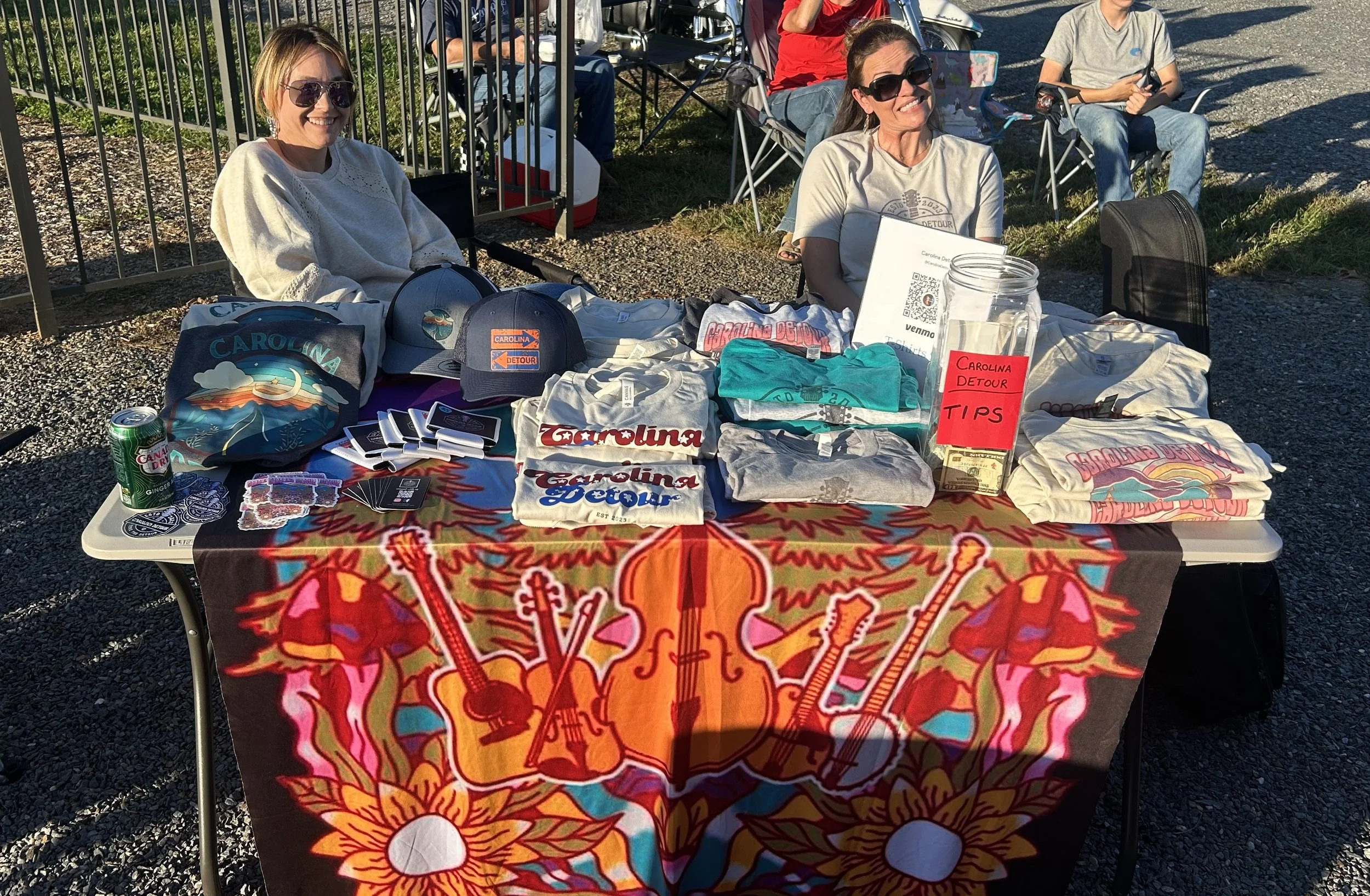 Two women sitting behind a table with Carolina Detour merchandise, including shirts, caps, and stickers, at an outdoor event. The table is covered with a colorful cloth featuring guitars and sunflowers. Other people are visible in the background on a
