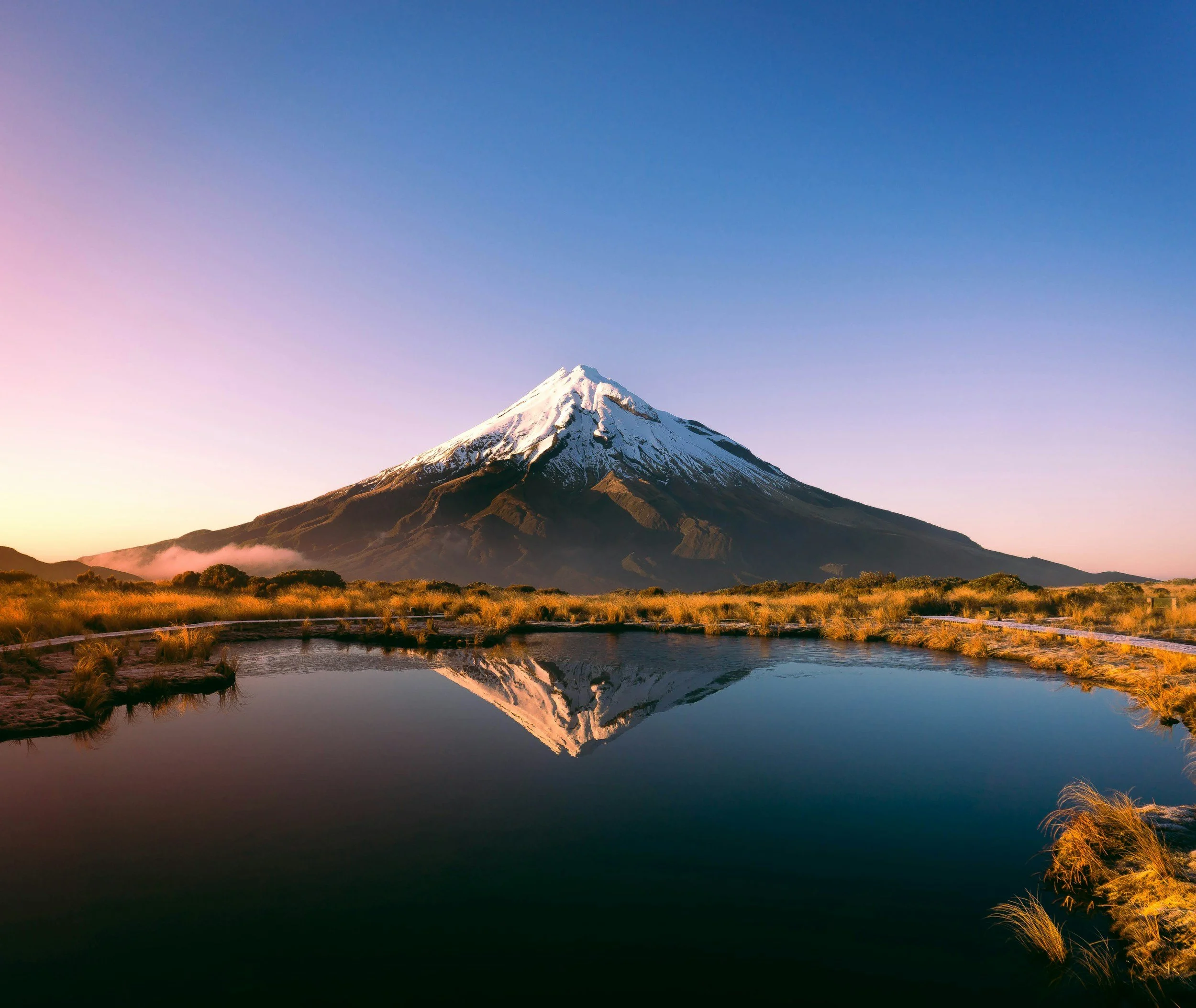 Snow-capped Mount Taranaki reflected in a calm lake at sunrise, with grassy landscape surrounding it.