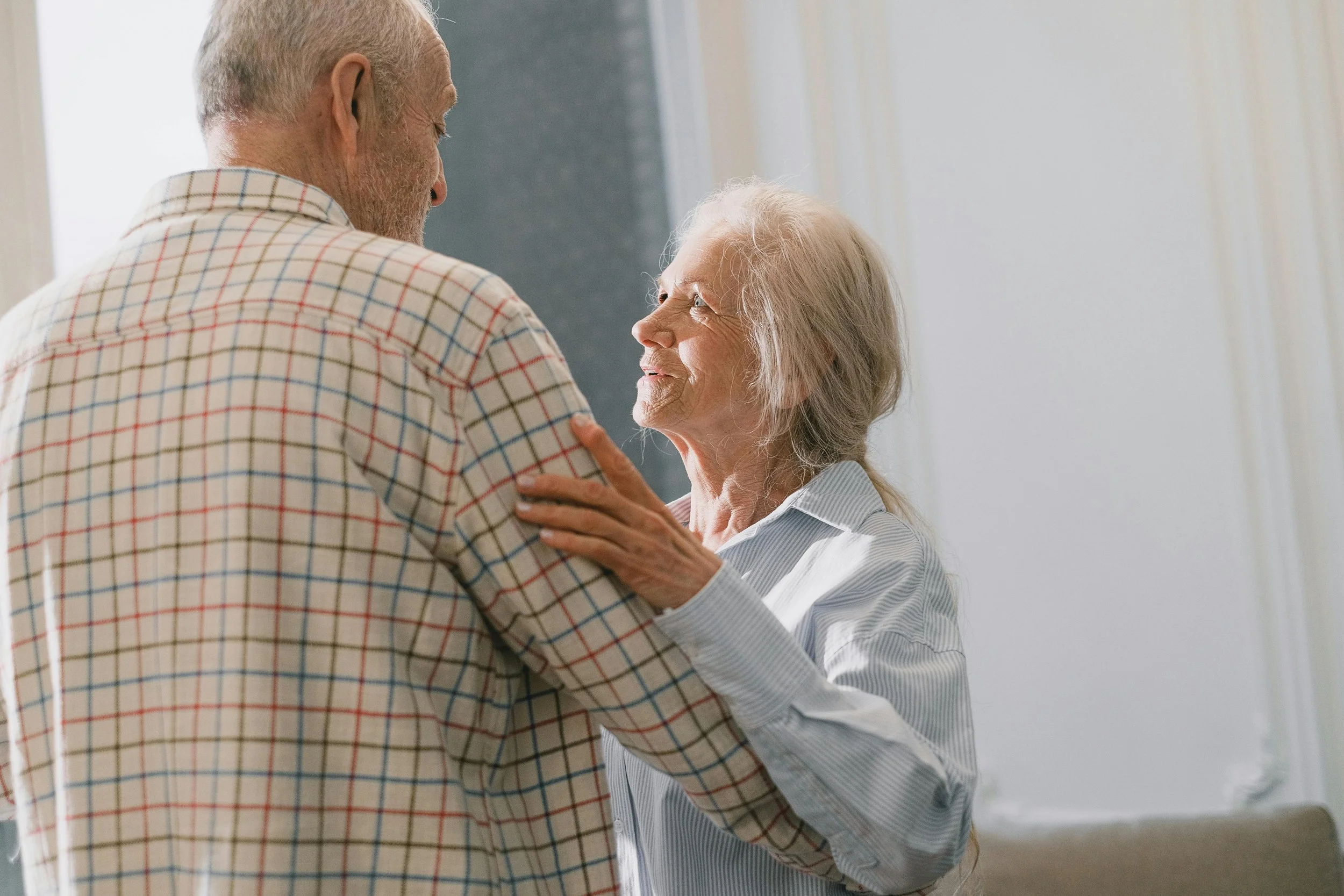 An elderly man and woman dancing and looking at each other happily in a room with natural light.