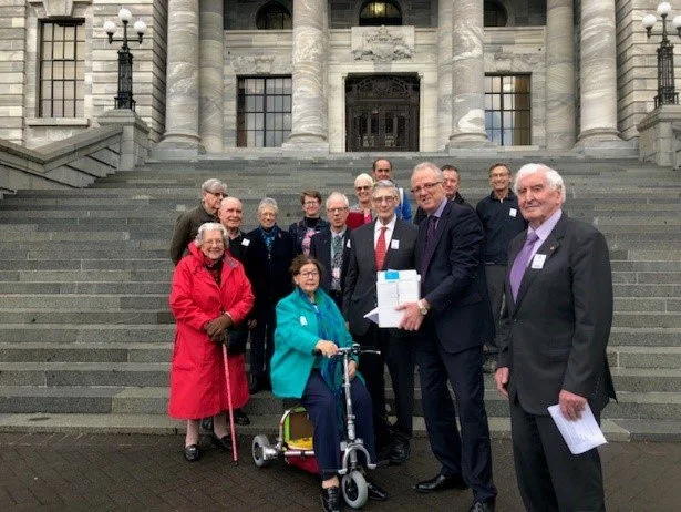 Group of people standing on outdoor steps in front of a historic building, with some holding papers and a woman using a mobility scooter.