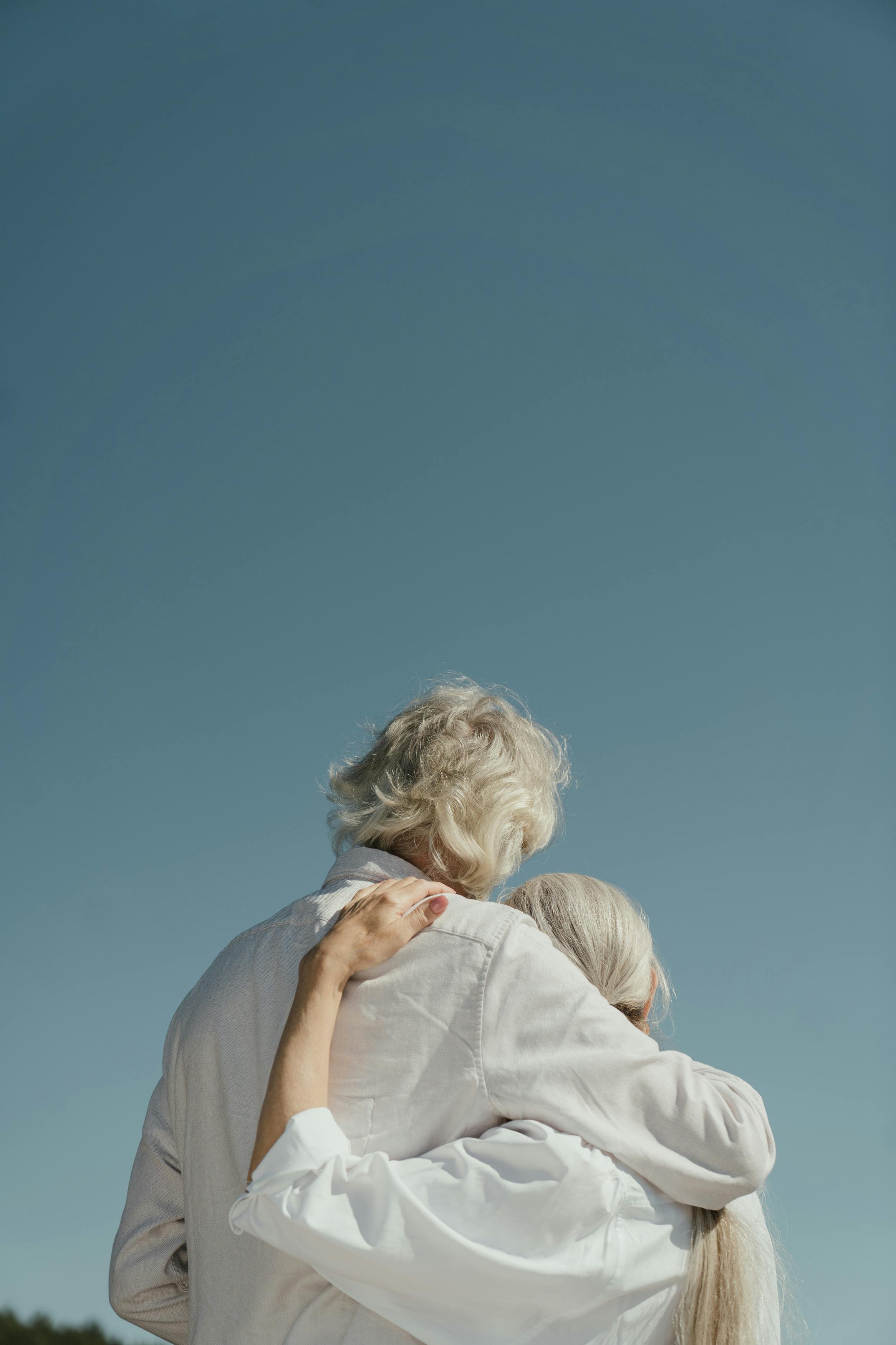 Two elderly women hugging outdoors under a clear blue sky.