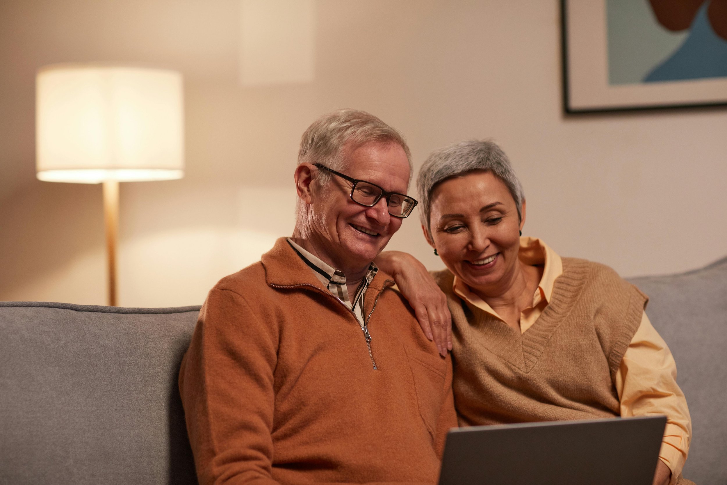 An elderly couple sitting on a couch smiling at a tablet, with a lamp and framed artwork in the background.