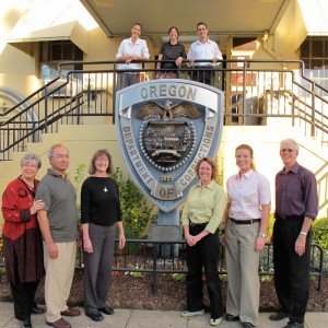 Group of ten people standing outdoors in front of the Oregon State Police badge plaque with a building behind them.
