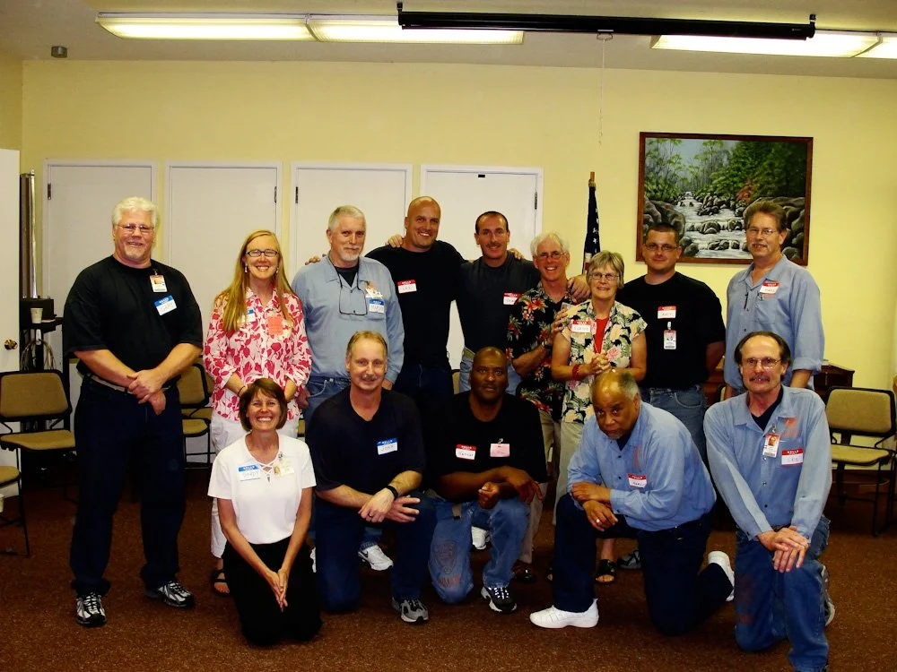 A group of 14 people, wearing name tags, posing for a photo in a room with beige walls and a painting of a river scene in the background. They are standing and kneeling in two rows.