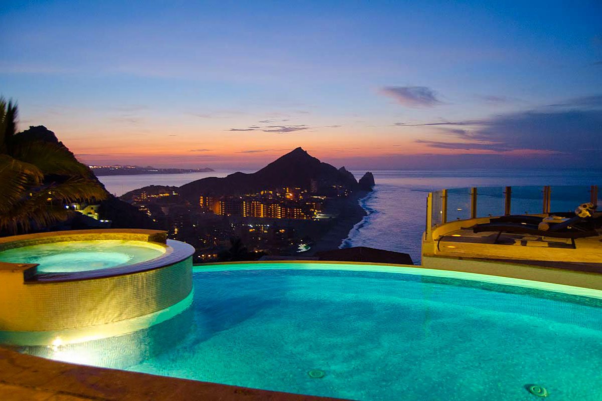 Infinity pool overlooking a coastal city at sunset with ocean view, mountains, and city lights in the distance.