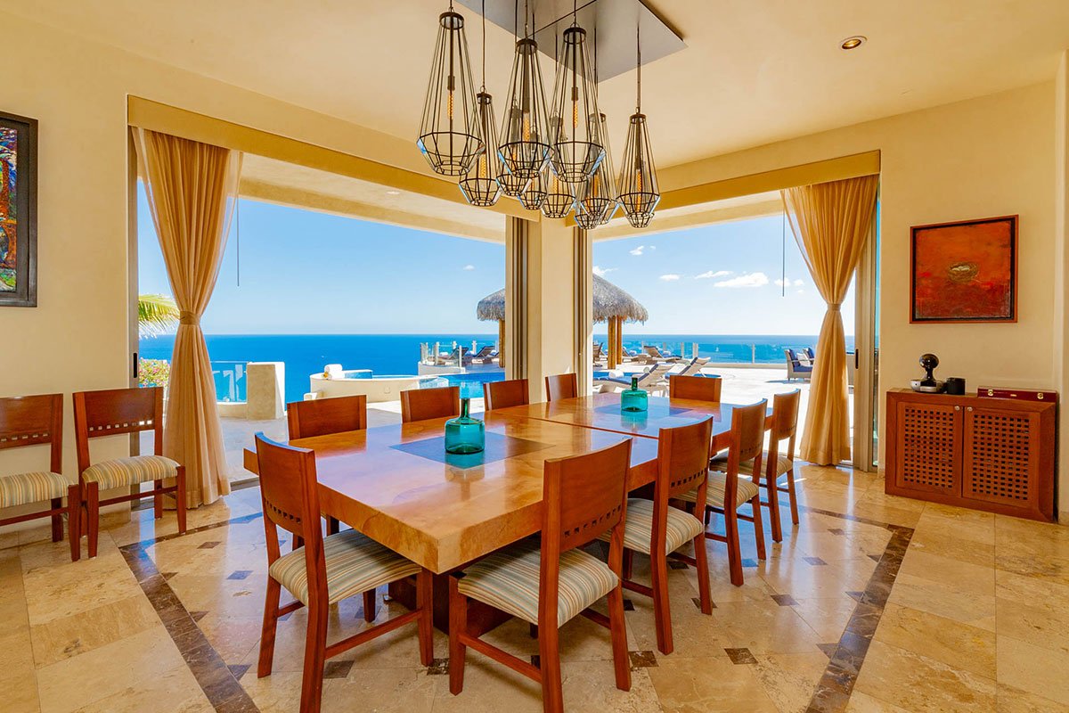 Luxurious dining area with wooden table and chairs, modern chandelier, and open view of an outdoor pool and ocean.