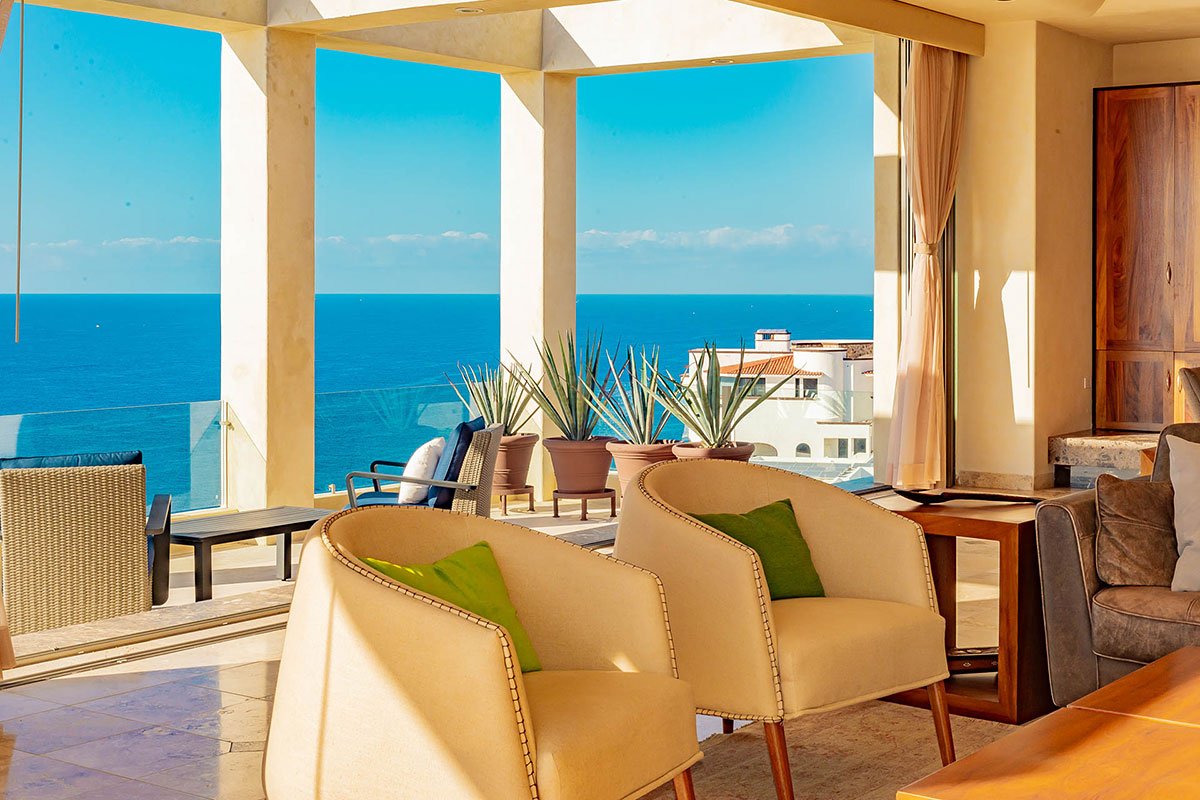 Luxury coastal living room with ocean view, beige chairs, and potted plants on a terrace.