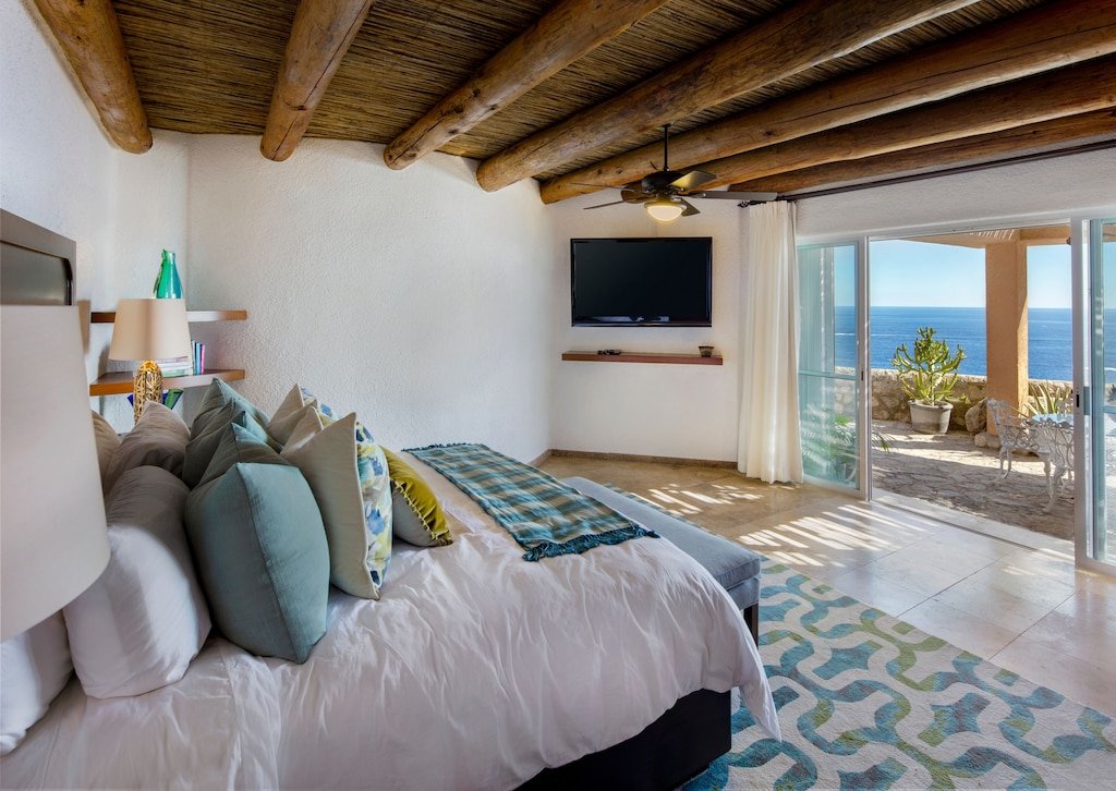 Luxurious bedroom with ocean view, featuring a large bed with multiple pillows, ceiling fan, wall-mounted TV, and sliding glass doors leading to a patio outside.