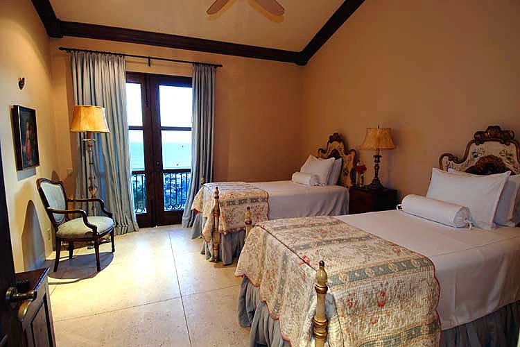 Cozy bedroom with two twin beds, decorative headboards, ornate bedding, and a side table with a lamp. A chair and a floor lamp are near a window with drawn curtains.
