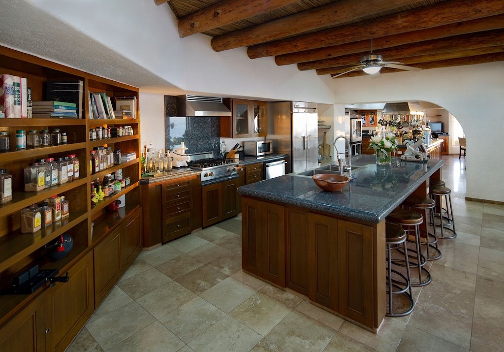 Spacious kitchen with wooden cabinets, large island with bar stools, open shelves with spices, stainless steel appliances, and exposed wooden beams on the ceiling.