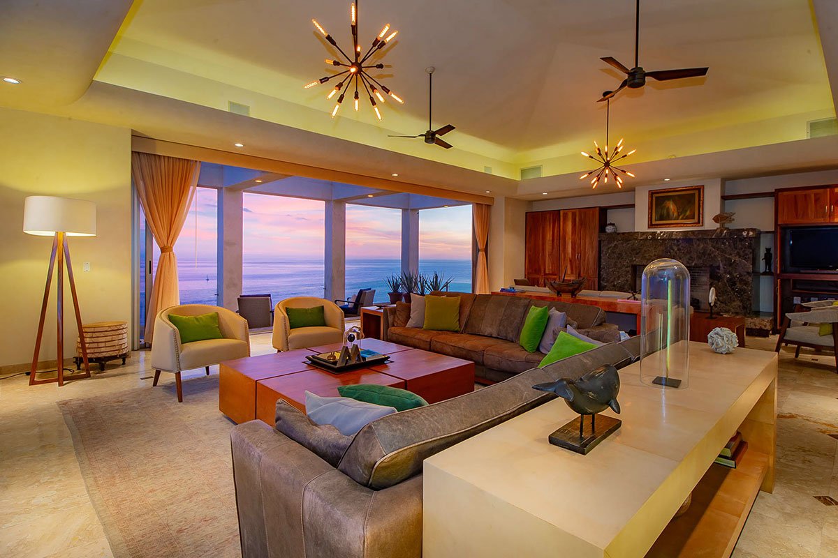 Luxury living room with modern decor, featuring a brown sectional sofa and green-and-brown accent chairs. Large windows offer an ocean view at sunset. Unique pendant lights and ceiling fans enhance the room's ambiance. A sculptural bird decorates the