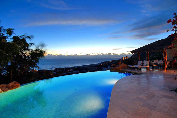 Infinity pool overlooking an ocean view at dusk, with a patio and outdoor seating area.