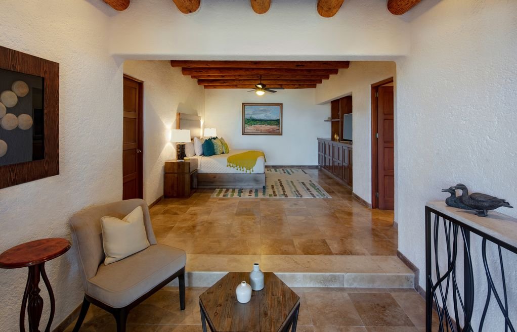 Cozy living room with a beige armchair, wooden coffee table, and decorative items. In the background, a bedroom with a bed, colorful rug, and artwork on the wall. Exposed wooden beams on the ceiling and tiled floor enhance the rustic aesthetic.