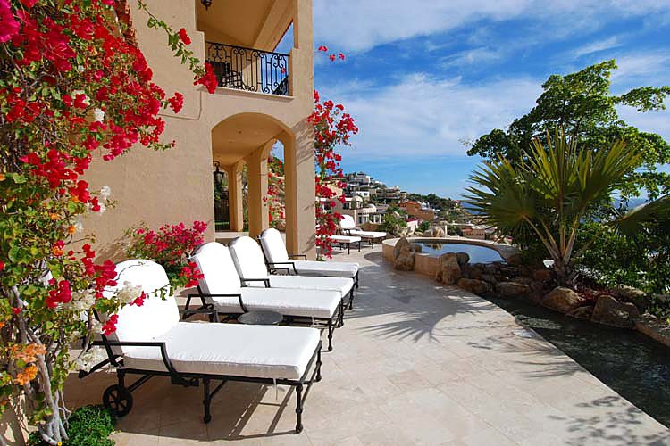 Luxury patio with white lounge chairs, bougainvillea, stone tiles, and a view of an infinity pool and distant hills.