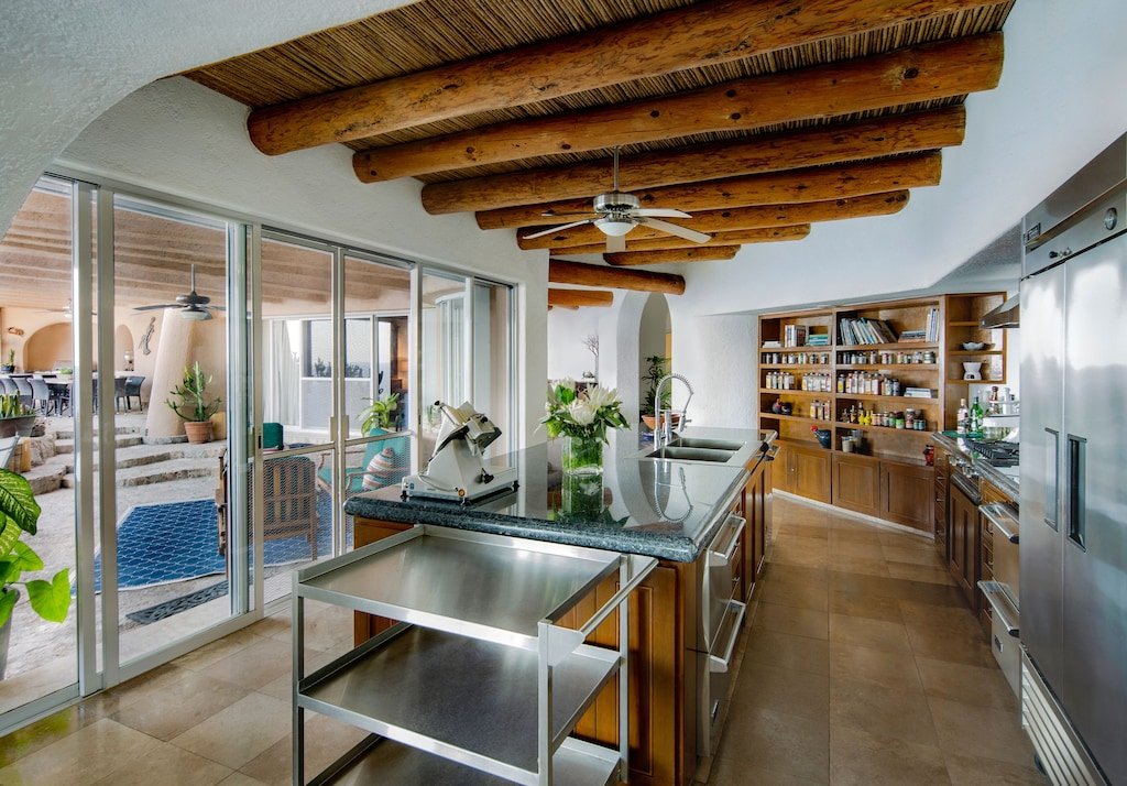 Modern kitchen featuring wooden beams, stainless steel appliances, an island with a sink, and open shelves with spices. Large windows reveal an outdoor patio area with plants and seating.