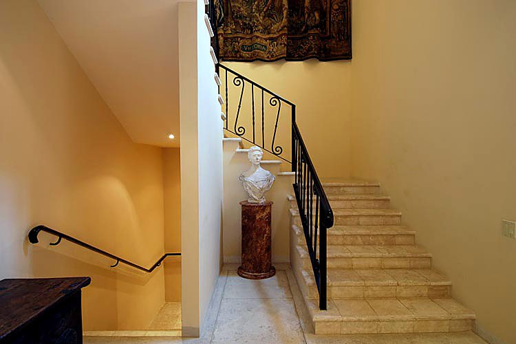 Interior staircase with tiled steps, ornate iron railing, marble bust, and tapestry on wall.