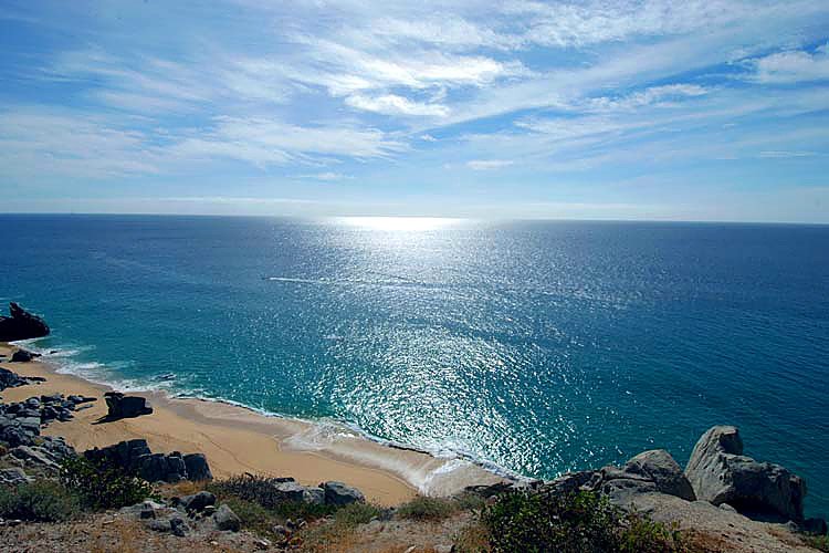 Scenic view of a beach with blue ocean and clear sky, waves lapping the sandy shore, surrounded by rocks and coastal vegetation.