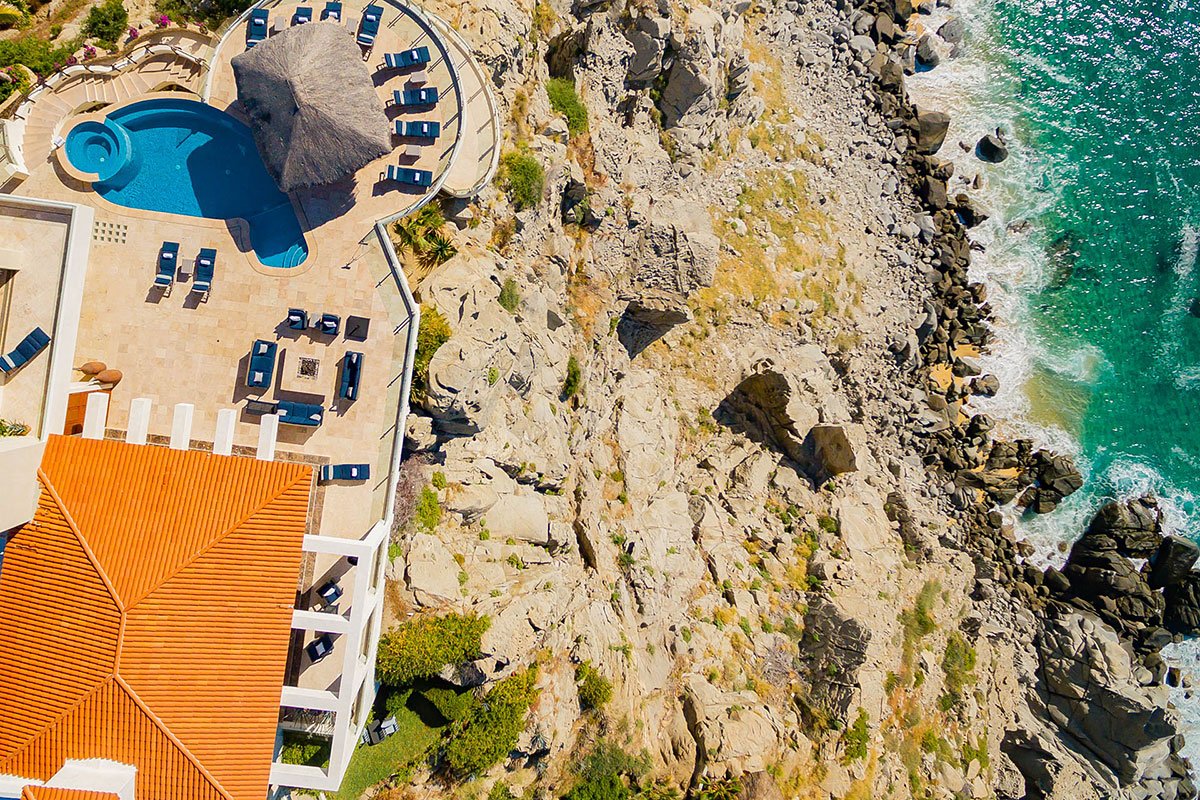 Aerial view of a luxury villa with a swimming pool, thatched-roof structure, sun loungers, and sitting area, situated on a rocky coastline next to the ocean.