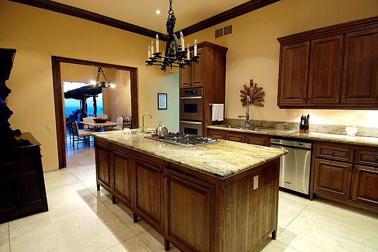 Modern kitchen interior with wooden cabinetry, granite countertops, a kitchen island, chandelier, stainless steel appliances, and a dining area visible through an open doorway.