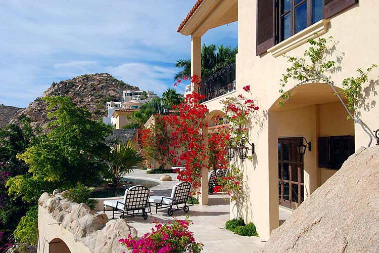 Mediterranean-style villa with red flowers, patio furniture, and hillside view.