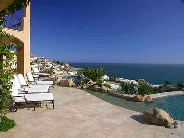 Luxury beachfront patio with lounge chairs, infinity pool, and ocean view under a clear blue sky.