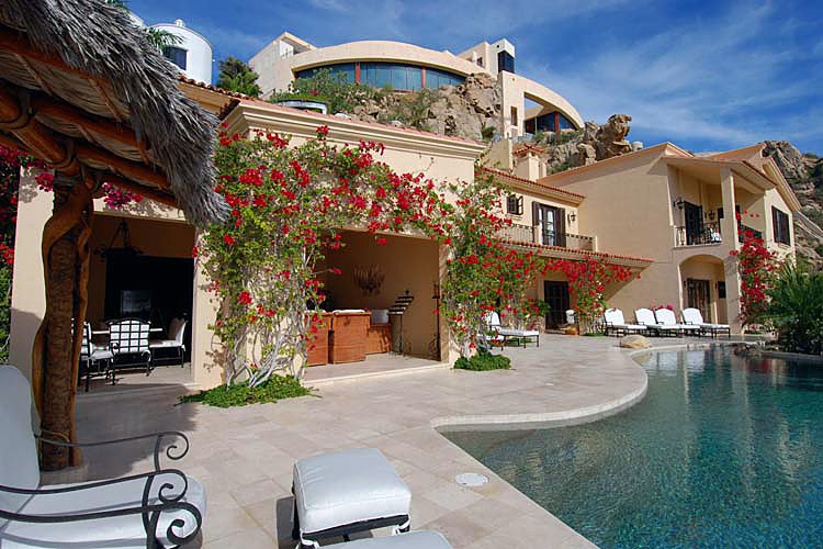 Luxurious villa with a large swimming pool and patio area, featuring outdoor seating and vibrant red bougainvillea climbing the walls, set against a rocky hillside under a blue sky.