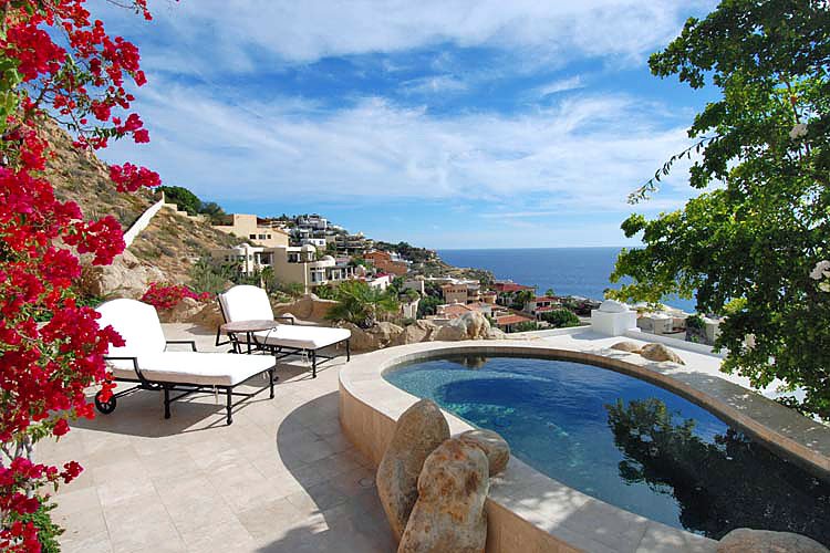 Luxury outdoor pool area with lounge chairs, overlooking ocean and hillside homes, surrounded by flowering plants and trees.