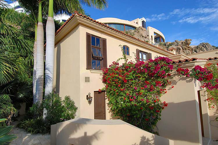 A Mediterranean-style house with beige walls, red roof tiles, and wooden shutters, surrounded by lush palm trees and vibrant bougainvillea. Clear blue sky and rocky terrain in the background.