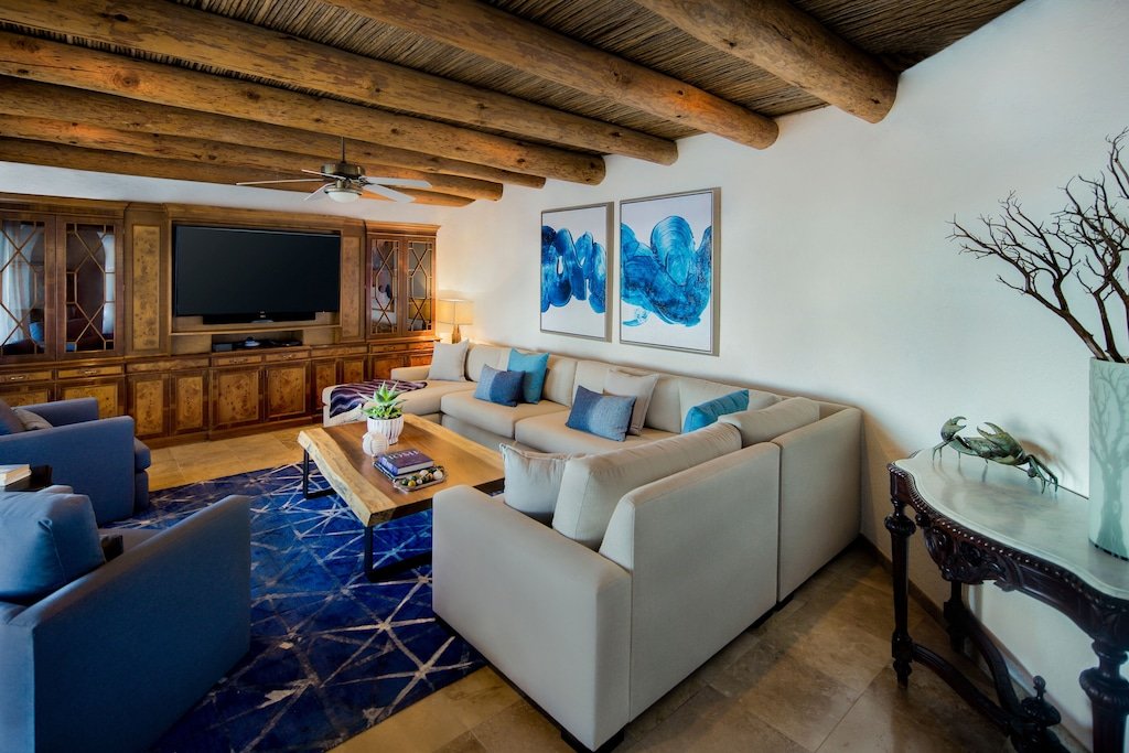 Cozy living room with beige sectional sofa, blue armchairs, wooden beamed ceiling, large TV, wooden cabinetry, blue abstract artwork, and decorative items.