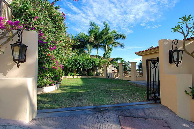 Gated entrance to a garden with palm trees and flowering plants, featuring black lantern-style lights and cream-colored walls under a blue sky with scattered clouds.