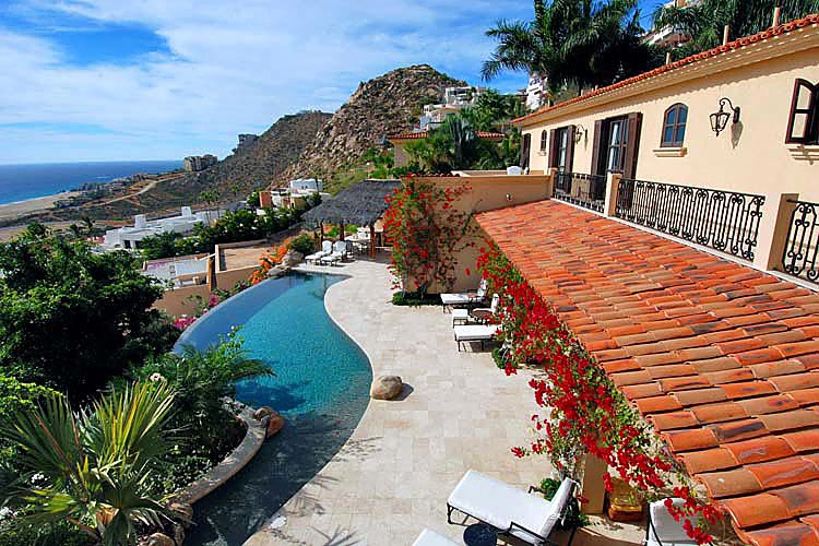 Luxury villa with infinity pool overlooking ocean and mountains, surrounded by lush plants and vibrant red flowers on terrace.
