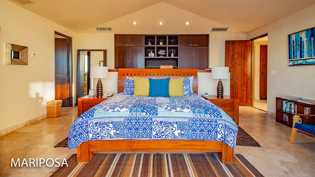 Spacious bedroom with a large wooden bed featuring blue and white patterned bedding, surrounded by modern decor including wooden nightstands with lamps, a colorful rug, and built-in shelving; "Mariposa" text in the corner.