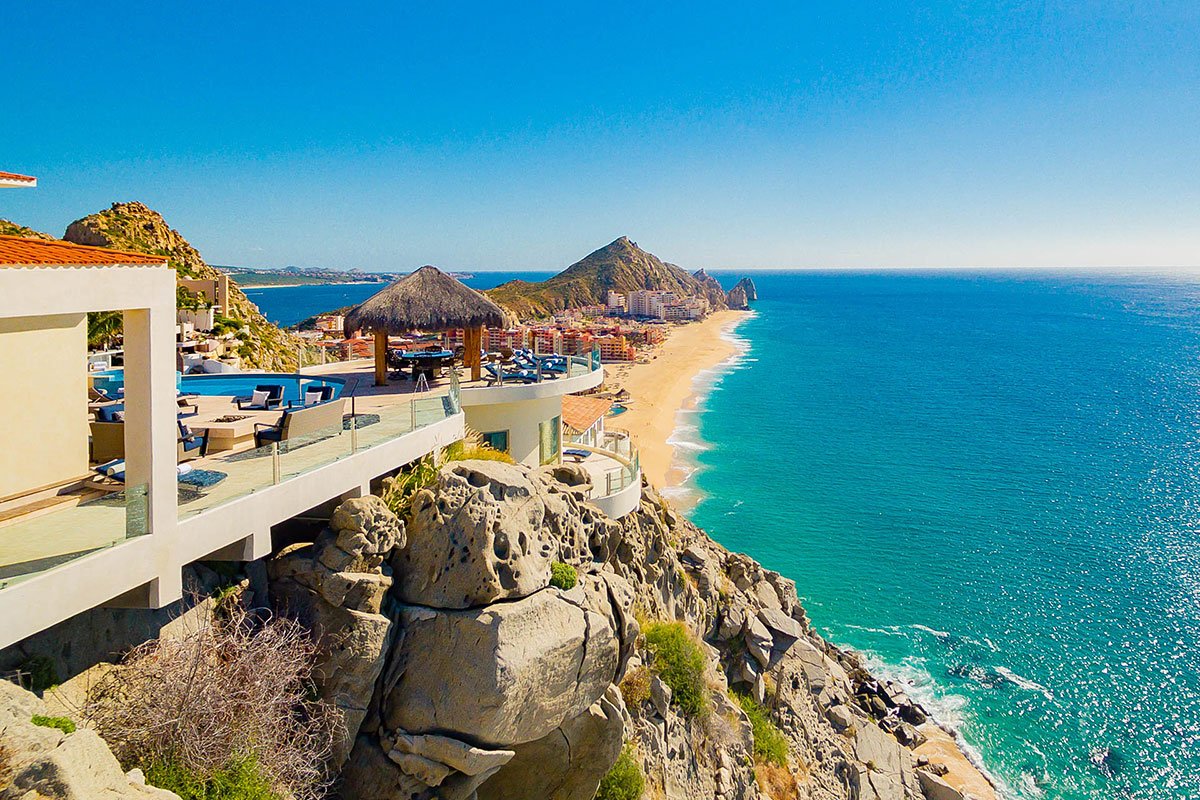 Scenic coastal view of a luxury villawith infinity pool, thatched-roof structures, and a sandy beach alongside the blue ocean.