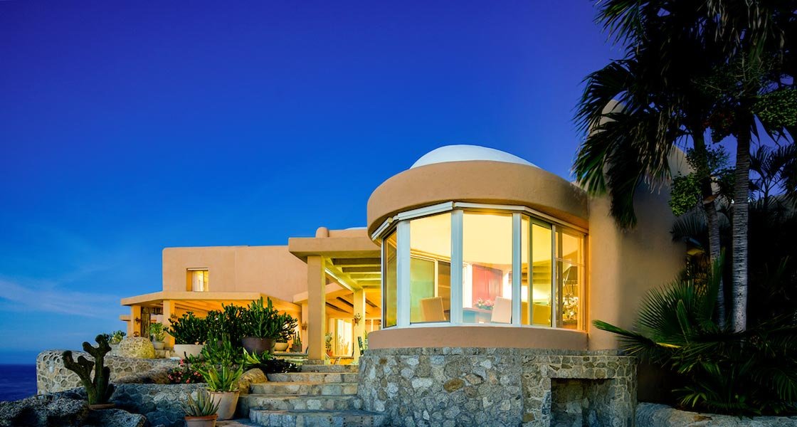 Modern villa with curved glass windows and stone exterior, surrounded by tropical plants, under a clear evening sky.