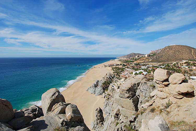 A coastal view of a sandy beach with rocky cliffs and blue ocean under a partly cloudy sky. In the distance, there are small buildings and hills.