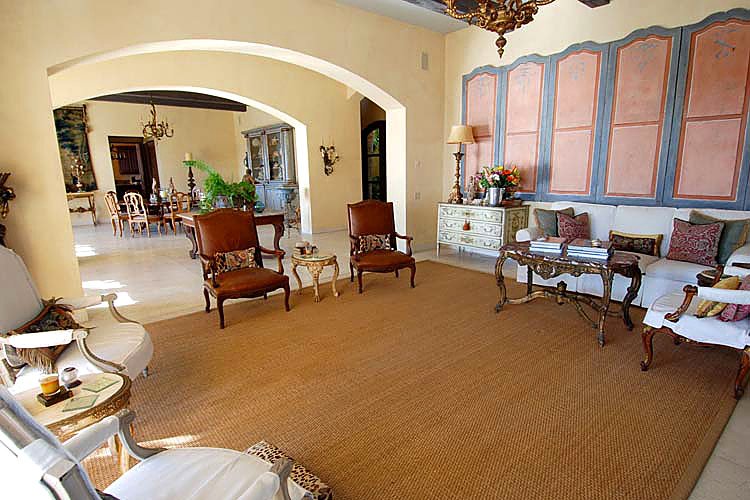 Elegant living room with classic furniture, featuring a large area rug, chairs, sofas, and decorative panels on the wall, leading into a dining room with a chandelier.