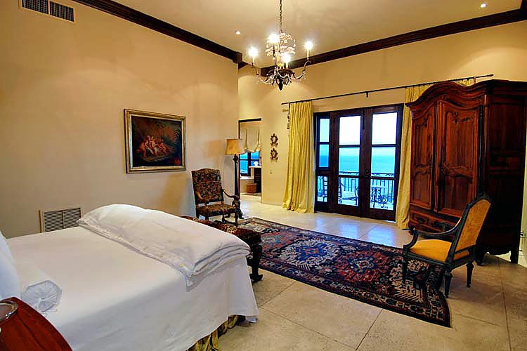Elegant bedroom with a large bed, ornate rug, wooden wardrobe, and a balcony view. The room is decorated with a painting and classical furniture. Yellow curtains frame the glass doors.