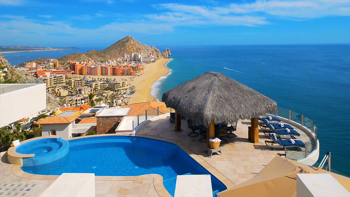 Luxury beachfront villa with a pool overlooking a sandy coastline and resort buildings, under a clear blue sky.