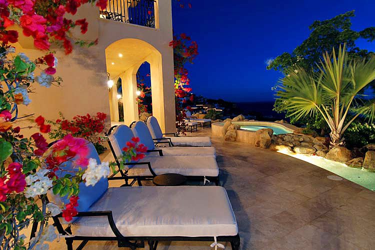 Luxury patio with lounge chairs and hot tub surrounded by tropical plants, illuminated at dusk.