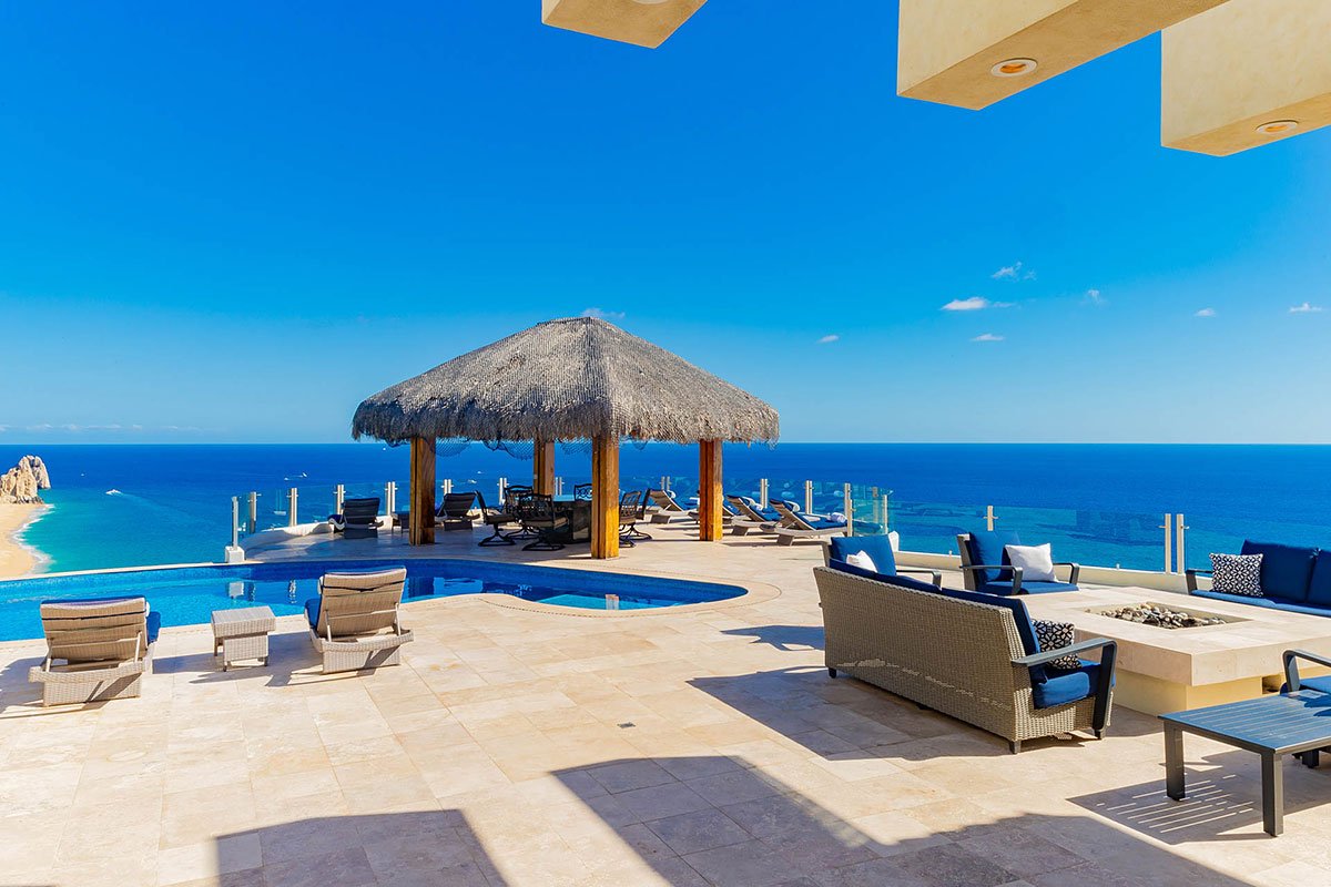 Luxury oceanfront terrace with infinity pool, sun loungers, fire pit seating, and palapa, overlooking a scenic beach.